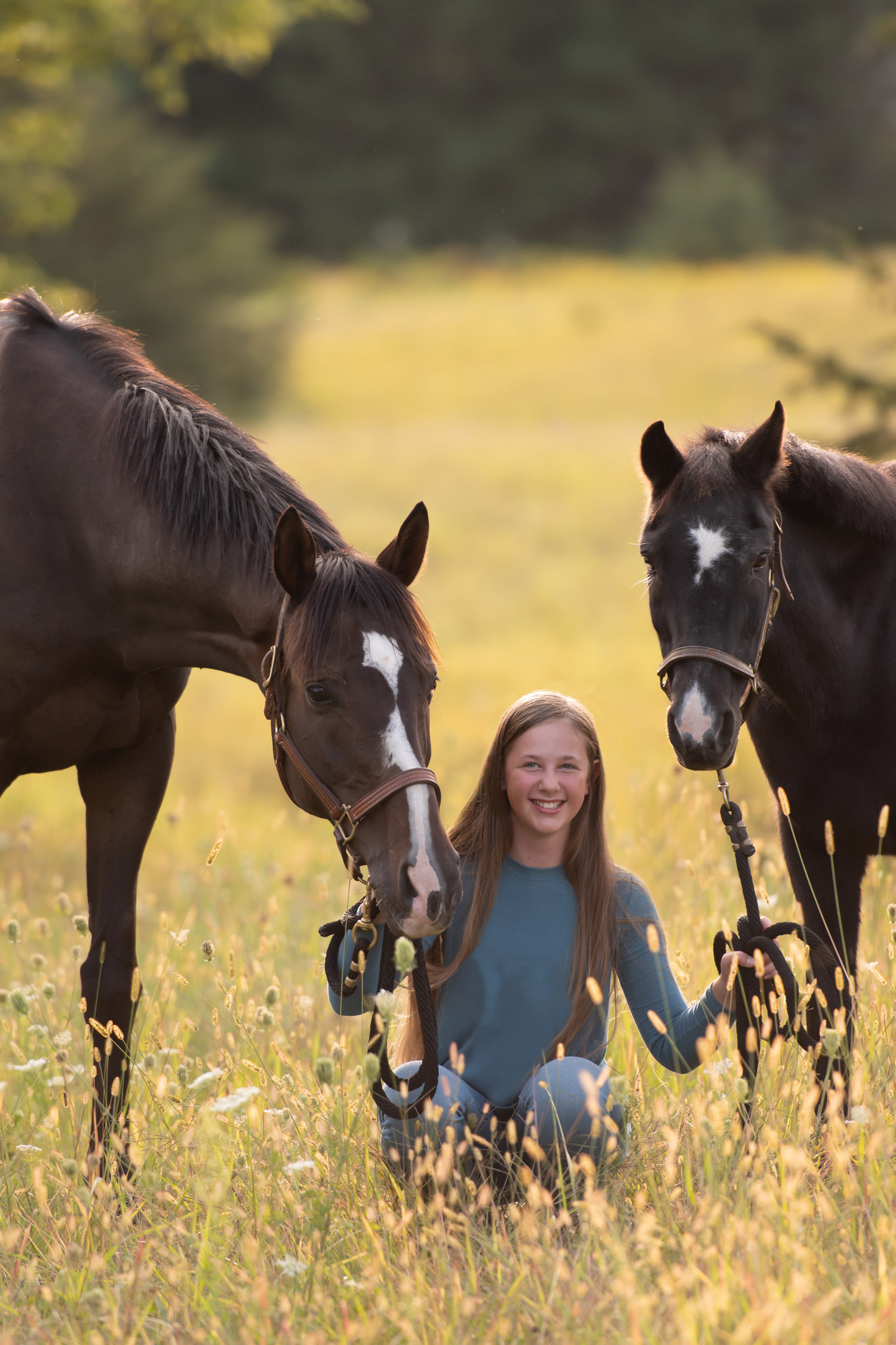 Isabella kneeling in a sunlit meadow between a Welsh pony and a black pony